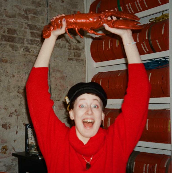 Analogue camera portrait of Vi Schreiber. They are wearing a bright red turtle neck pullover and holding the plastic model of a red lobster over their head. They are looking excitedly into the camera, as if screaming from joy.